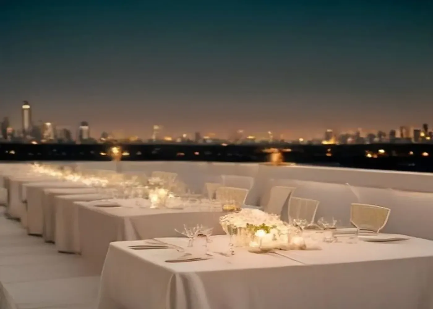 A long candlelit dinner table with white flowers on the White House rooftop at dusk, the city skyline beyond